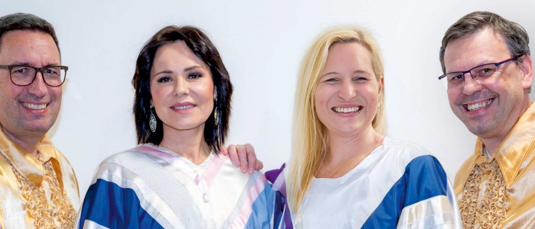 Vier Menschen stehen eng für ein Foto zusammen. Zwei Frauen tragen weiße und blaue Roben mit weißen Umhängen. Der Mann rechts trägt eine Brille und ein goldenes Hemd.
