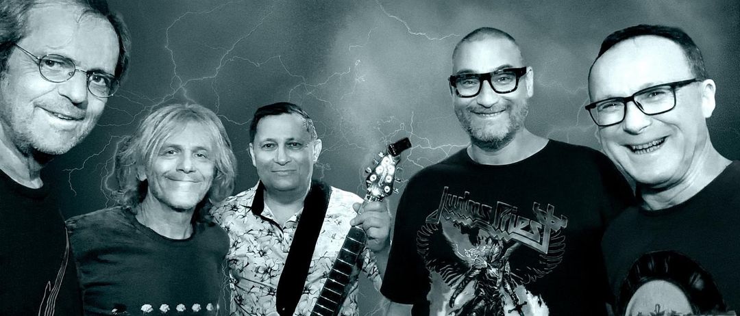 Eine Band namens CM2GTHR mit vier Mitgliedern steht vor einem dunklen Hintergrund mit Blitzen. Sie alle haben lächelnde Gesichter.