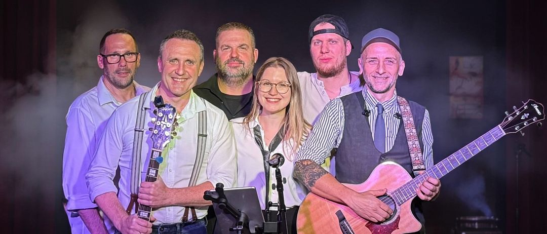 Eine Band mit sechs Mitgliedern steht lächelnd zusammen. Sie halten Gitarren, und einige haben Mikrofone. Die Frau in der Mitte trägt eine Brille. Der Mann auf der rechten Seite trägt eine Krawatte.