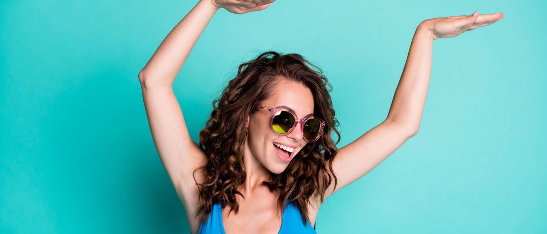 Eine Frau mit lockigem Haar in einem blauen Tanktop und Sonnenbrille steht mit ausgestreckten Armen und lächelt vor einem hellblauen Hintergrund.