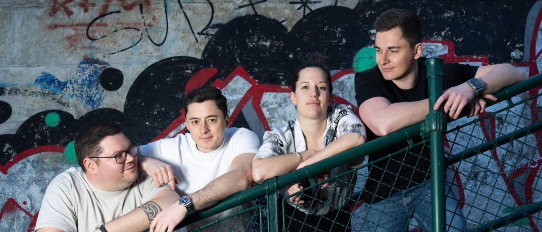 Vier Personen stehen gemeinsam auf einer Treppe mit Graffiti, lächelnd und posieren für ein Foto. Die Geländer sind grün und die Wand hinter ihnen hat bunte Graffiti.