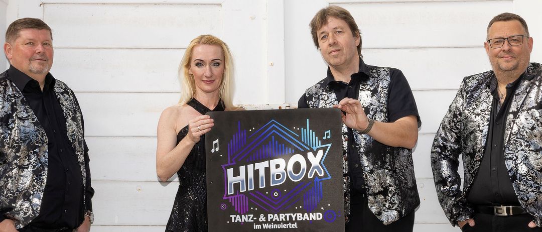 Eine Gruppe von vier Musikern, zwei Männer und zwei Frauen, stehen eng beieinander. Sie sind in glänzenden Anzügen gekleidet und halten ein Plakat mit der Aufschrift HITBOX. Sie stehen vor einer weißen Wand.