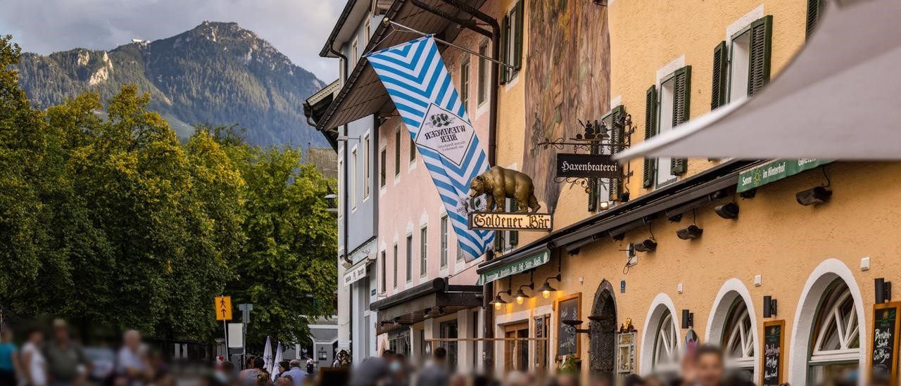 Eine überfüllte Straße mit Gebäuden, die eine blau-weiße Flagge, einen goldenen Bären und ein Schild für Goldener Bar zeigen. Im Hintergrund befindet sich eine Bergkette unter einem teilweise bewölkten Himmel.