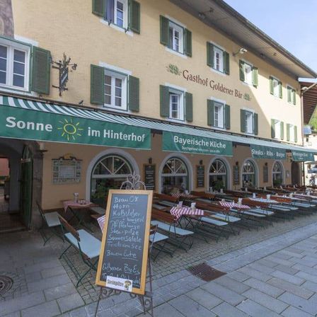 Ein Restaurant mit grünem Vordach, das ein Kreidetafelmenü und Sitzgelegenheiten im Freien mit Tischen und Stühlen bietet. Das Schild lautet 'Sonne im Hinterhof'.