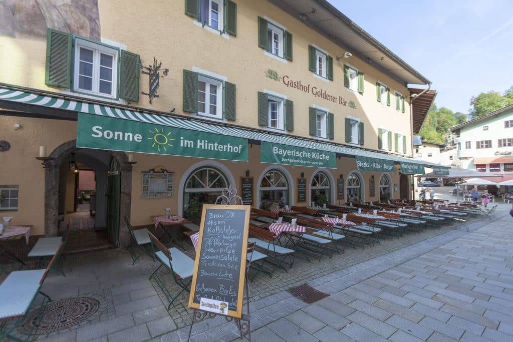 Ein Restaurant mit grünem Vordach, das ein Kreidetafelmenü und Sitzgelegenheiten im Freien mit Tischen und Stühlen bietet. Das Schild lautet 'Sonne im Hinterhof'.