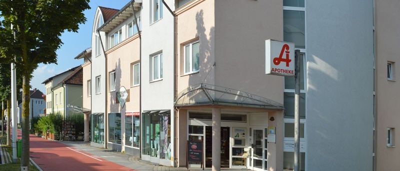 Ein Apothekengebäude mit einem roten Buchstaben A auf einem Schild steht an einer Straße. Das Gebäude besteht aus hellen Ziegeln und Glasfenstern. Auf der linken Seite des Gebäudes befindet sich ein Baum.
