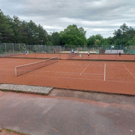 Ein Tennisplatz mit rotem Sandplatz. Mehrere Spieler in blauen und weißen Uniformen üben. Ein Metallzaun umgibt den Platz. Bäume und ein bewölkter Himmel im Hintergrund.