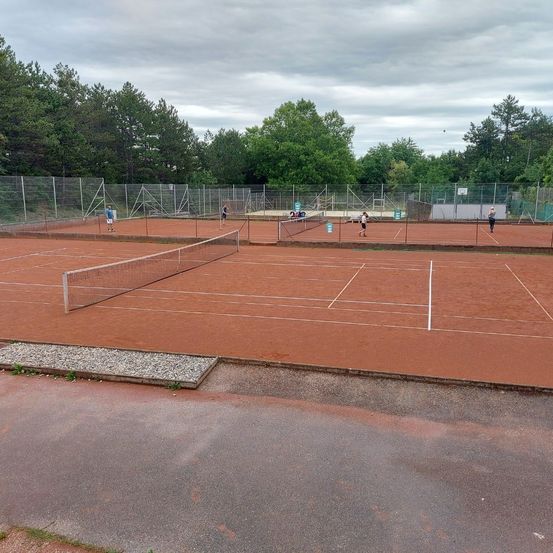 Ein Tennisplatz mit rotem Sandplatz. Mehrere Spieler in blauen und weißen Uniformen üben. Ein Metallzaun umgibt den Platz. Bäume und ein bewölkter Himmel im Hintergrund.