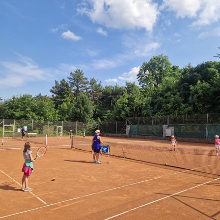 An einem sonnigen Tag spielen mehrere junge Menschen Tennis auf Sandplätzen, umgeben von Bäumen und einem klaren blauen Himmel.