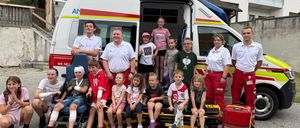 Eine Gruppe von Kindern und Erwachsenen posiert für ein Foto vor einem Krankenwagen, wobei einige Kinder auf einer Trage sitzen und andere hinter dem Krankenwagen stehen.
