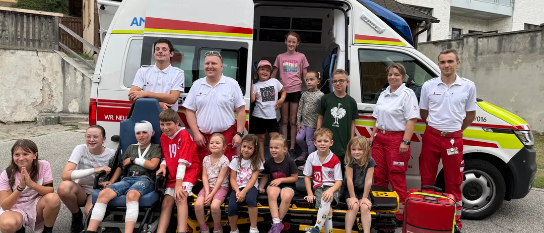 Eine Gruppe von Kindern und Erwachsenen posiert für ein Foto vor einem Krankenwagen, wobei einige Kinder auf einer Trage sitzen und andere hinter dem Krankenwagen stehen.