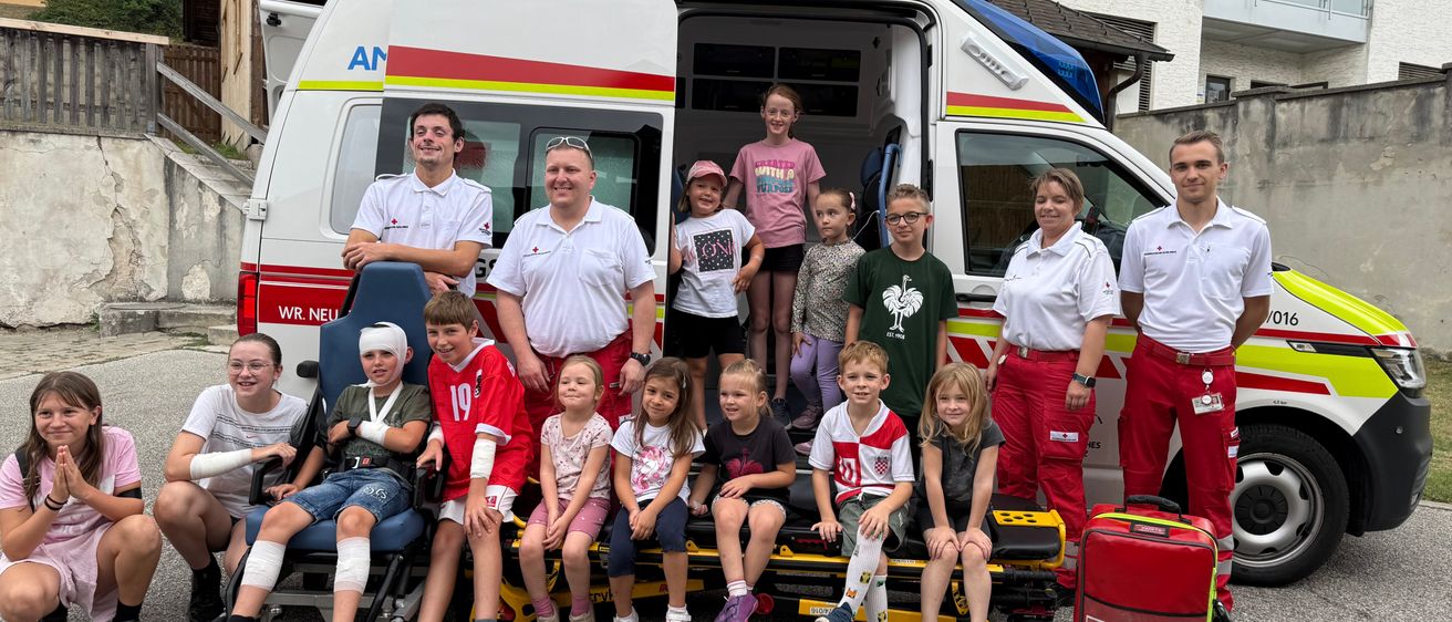 Eine Gruppe von Kindern und Erwachsenen posiert für ein Foto vor einem Krankenwagen, wobei einige Kinder auf einer Trage sitzen und andere hinter dem Krankenwagen stehen.