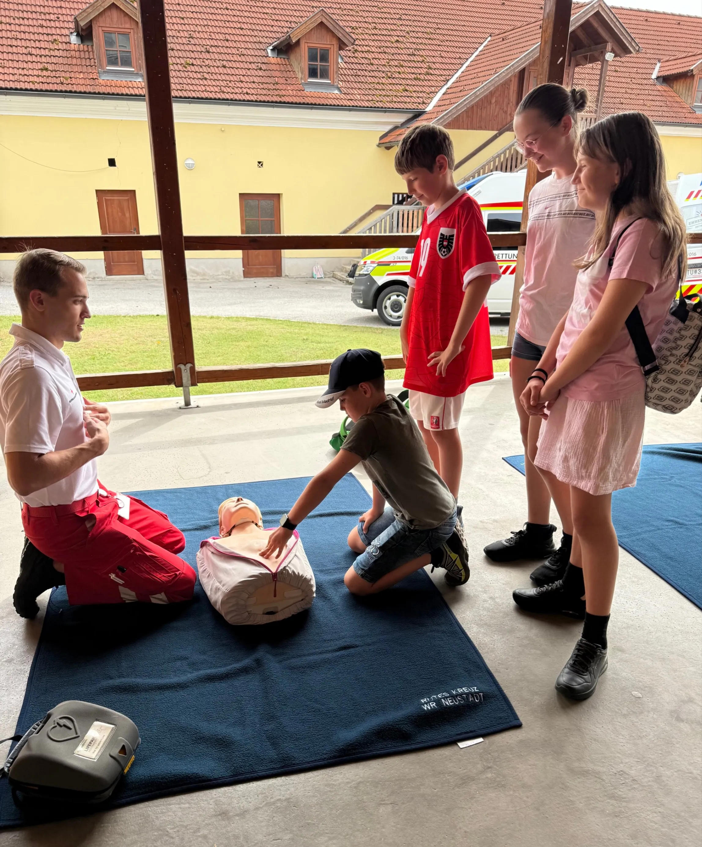 Eine Gruppe von Kindern und Erwachsenen ist um einen CPR-Trainingspuppen herum versammelt, die auf einem blauen Teppich liegt. Ein Erwachsener kniet neben der Puppe, während ein anderer dahinter steht, und zwei Kinder beobachten. Der Ort scheint ein Außentrainingsbereich mit einem Gebäude im Hintergrund zu sein.