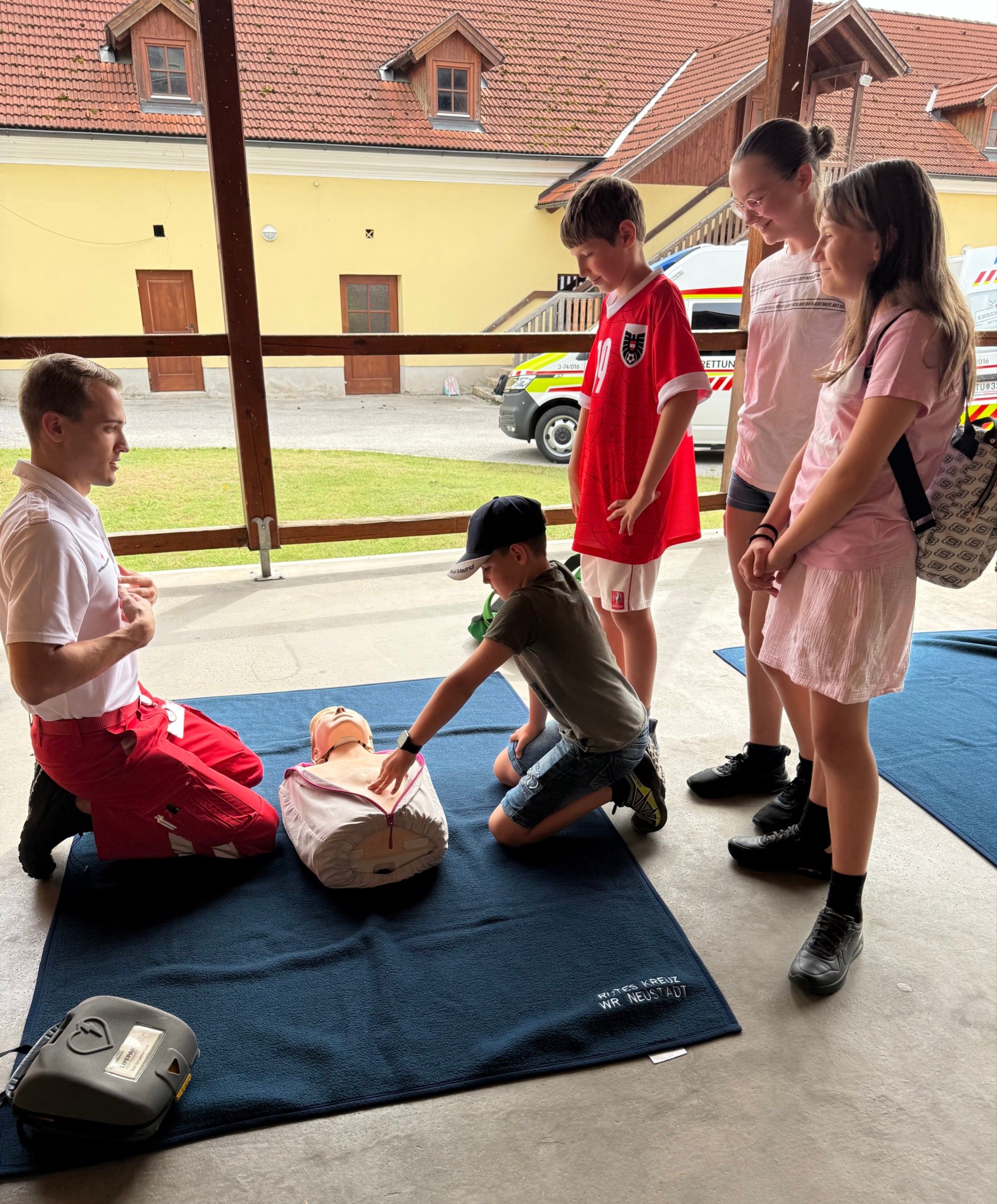 Eine Gruppe von Kindern und Erwachsenen ist um einen CPR-Trainingspuppen herum versammelt, die auf einem blauen Teppich liegt. Ein Erwachsener kniet neben der Puppe, während ein anderer dahinter steht, und zwei Kinder beobachten. Der Ort scheint ein Außentrainingsbereich mit einem Gebäude im Hintergrund zu sein.