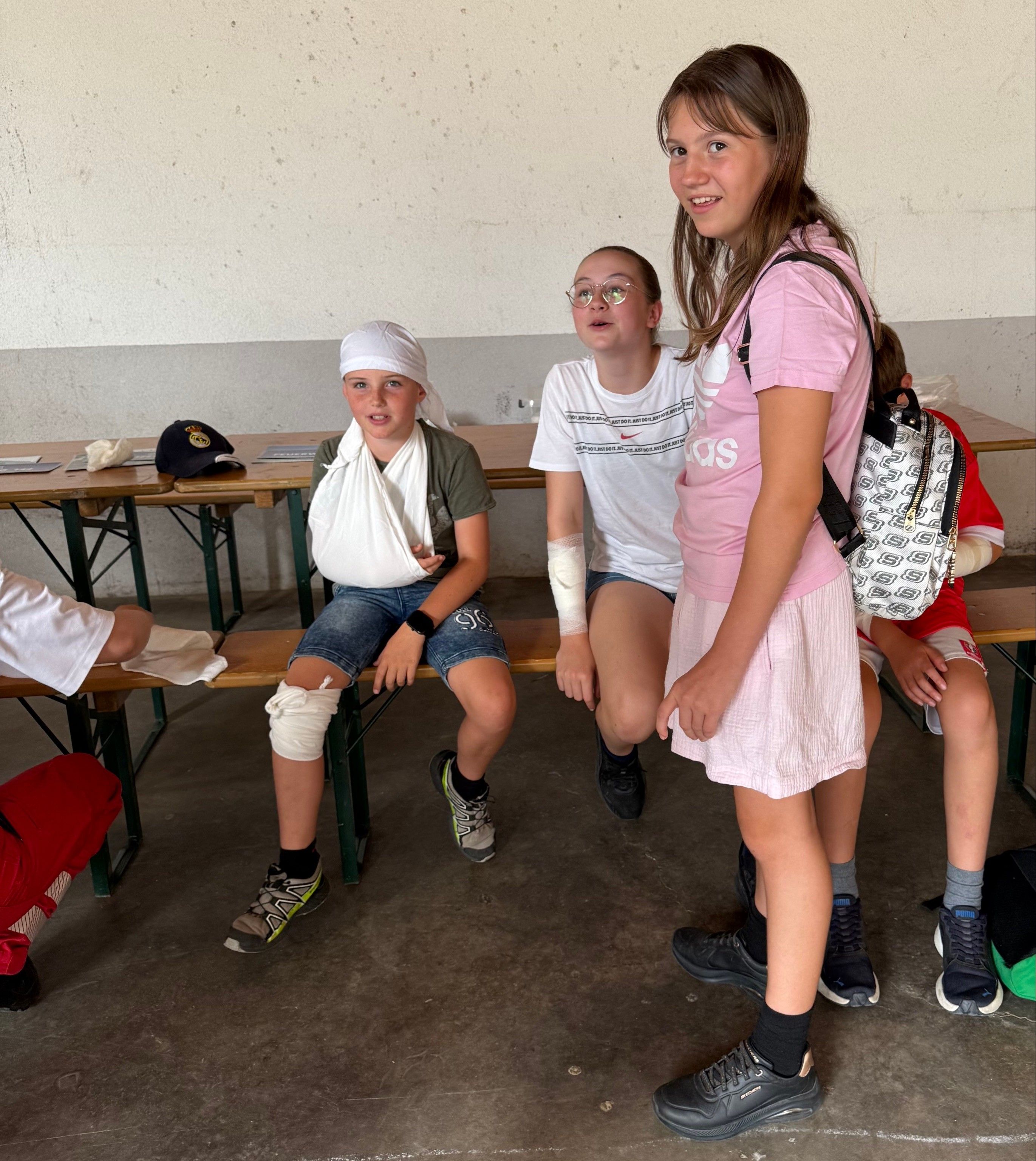 Drei Kinder sitzen auf einer Bank. Eines hat einen bandagierten Arm, ein anderes einen bandagierten Fuß, und das dritte trägt eine Brille. Ein Mädchen mit einer Tasche steht in der Nähe.
