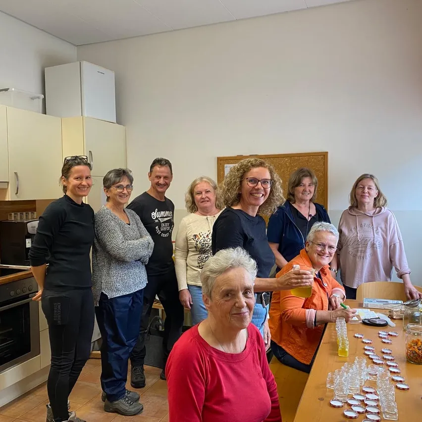 Eine Gruppe von Menschen steht in einer Küche, lächelt und posiert für ein Foto. Sie sind von Flaschen, Gläsern und einem Tisch mit kleinen Glasbehältern umgeben. Eine Frau in einem orangefarbenen Shirt hält eine Flasche und scheint etwas einzuschenken.