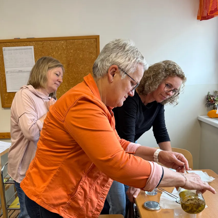 Drei Frauen sind in einem Raum, zwei von ihnen schauen in einen Glaskrug, und eine lächelt. Die Frau in der orangefarbenen Jacke hält den Krug. Vor ihnen steht ein Tisch mit einem Krug und einer Flasche.
