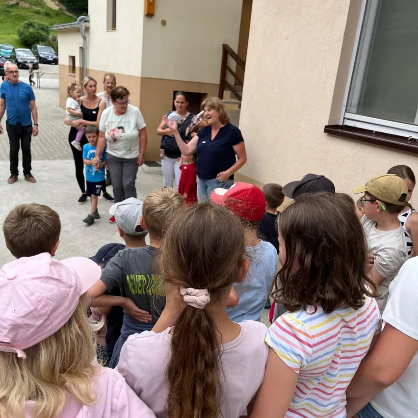 Eine Gruppe von Menschen, einschließlich Erwachsener und Kindern, steht vor einem Gebäude. Eine Frau hält eine Flasche und lächelt. Einige Kinder tragen Mützen.