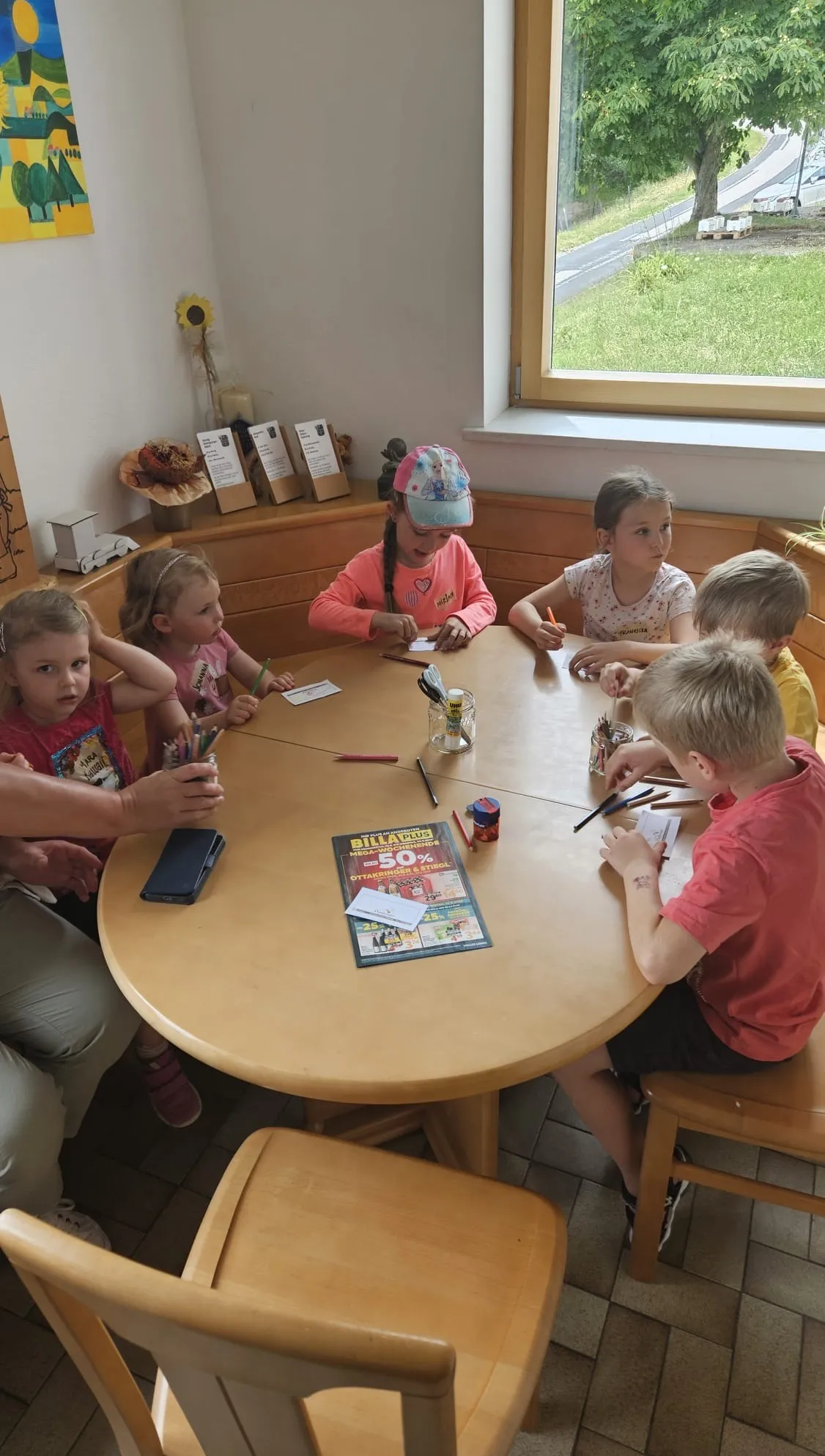 Eine Gruppe von Kindern sitzt um einen Tisch herum und malt. Eines von ihnen trägt einen Hut. Sie sind in einem Raum mit einem Fenster, das ein Grasfeld zeigt.