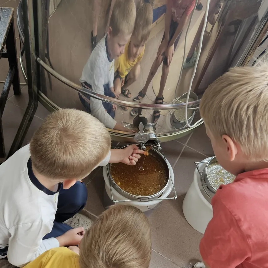 Vier Jungen sind um einen großen Metallbehälter versammelt, möglicherweise bei einer Koch- oder Brauaktivität. Ein Junge gießt eine Flüssigkeit in den Behälter.