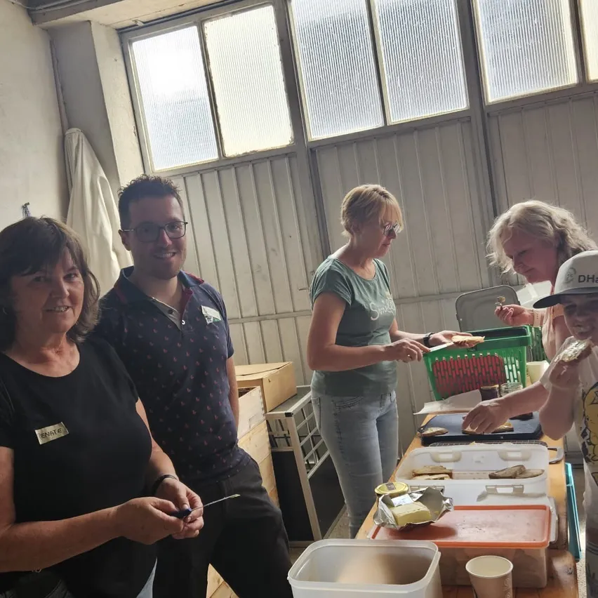 Eine Gruppe von Menschen versammelt sich in einem Raum, möglicherweise zur Essenszubereitung. Eine Frau hält ein Sandwich, und eine andere hält einen Tablett mit Essen. Ein Mann steht hinter ihnen, und eine Frau steht neben ihm. Es gibt Lagerboxen und einen Schreibtisch im Raum.