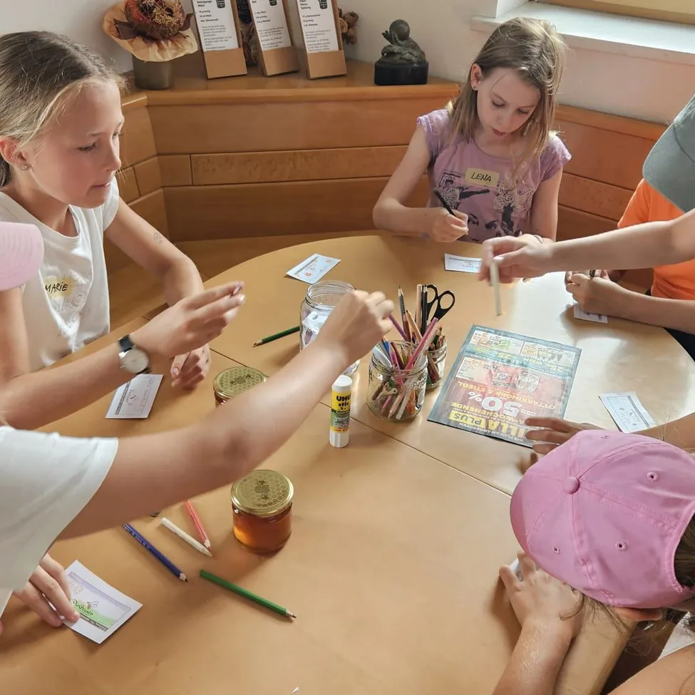 Eine Gruppe von Kindern sitzt um einen Tisch herum und ist in einer Bastelaktivität beschäftigt. Sie haben verschiedene Bastelutensilien wie Stifte, Scheren und Gläser mit Kleber. Ein Kind hält ein Stück Papier und einen Stift.