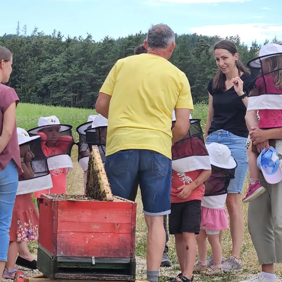 Eine Gruppe von Menschen, einschließlich Kindern, steht um einen roten Bienenstock auf einem Feld und trägt Schutzschleier. Ein Mann in einem gelben T-Shirt und eine Frau in einem schwarzen T-Shirt stehen vor dem Bienenstock.