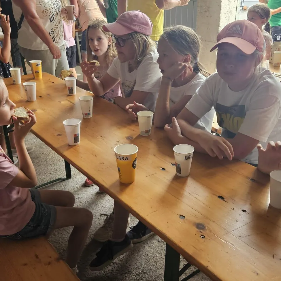 Fünf Kinder sitzen an einem Holztisch und essen Sandwiches, mit Tassen und Tellern vor ihnen. Ein paar Erwachsene stehen hinter ihnen, und einer hält eine Tasse.