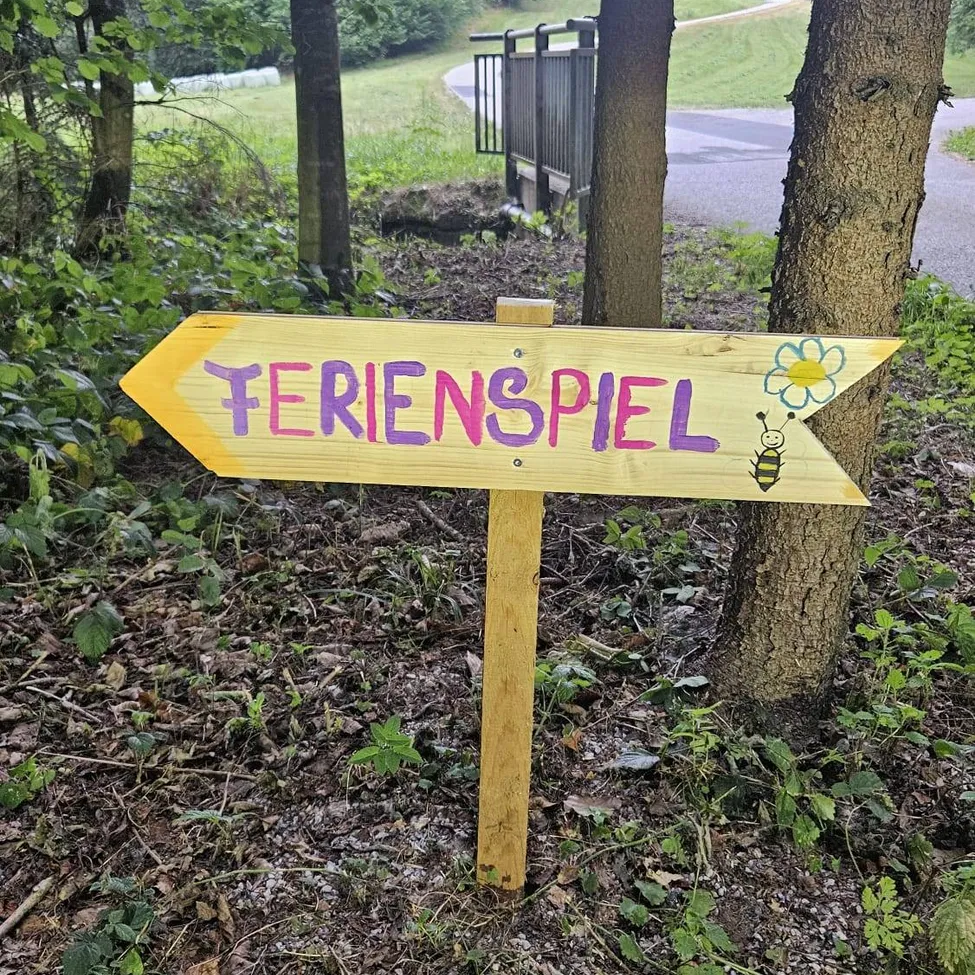 Ein Holzschild mit dem Wort Ferienspiel in bunten Buchstaben und einer Bienenillustration auf einem gelben Pfeil, der nach rechts zeigt, in einer natürlichen Umgebung mit Bäumen und Pflanzen platziert.