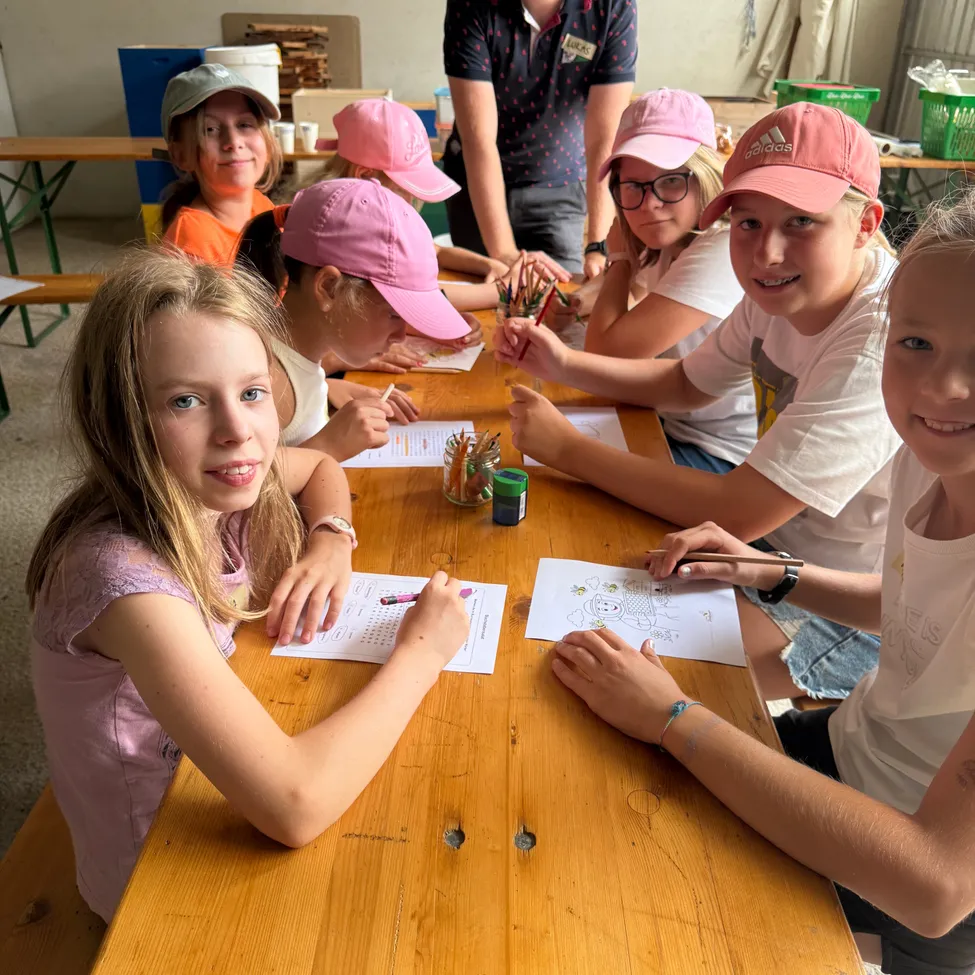 Fünf junge Mädchen mit rosa Mützen sitzen an einem Tisch und beschäftigen sich mit Malaktivitäten. Sie sind von Malbüchern, Stiften und Markern umgeben. Ein Mann steht hinter ihnen und beaufsichtigt die Aktivität.