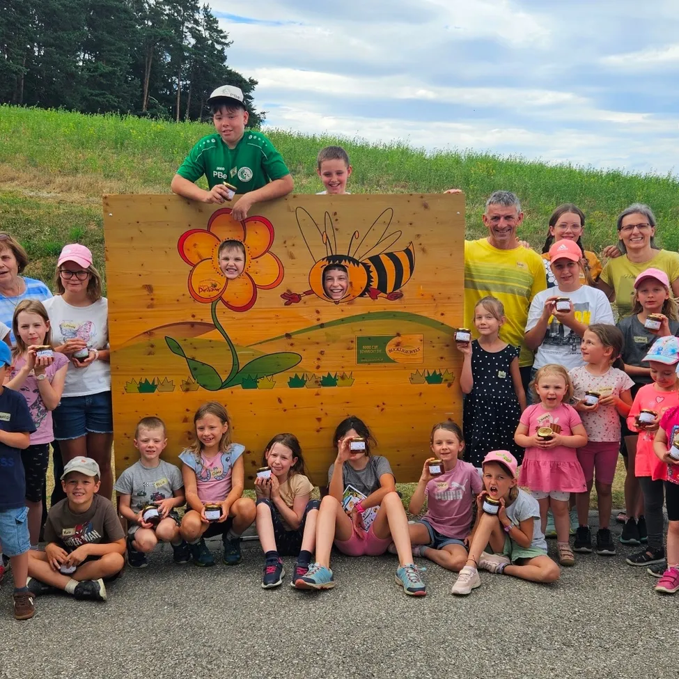 Eine Gruppe von Kindern und Erwachsenen posiert für ein Foto vor einem hölzernen Schild mit Bienen- und Blumendesign und hält kleine Gläser. Im Hintergrund befindet sich eine grasbewachsene Anhöhe und ein bewölkter Himmel.