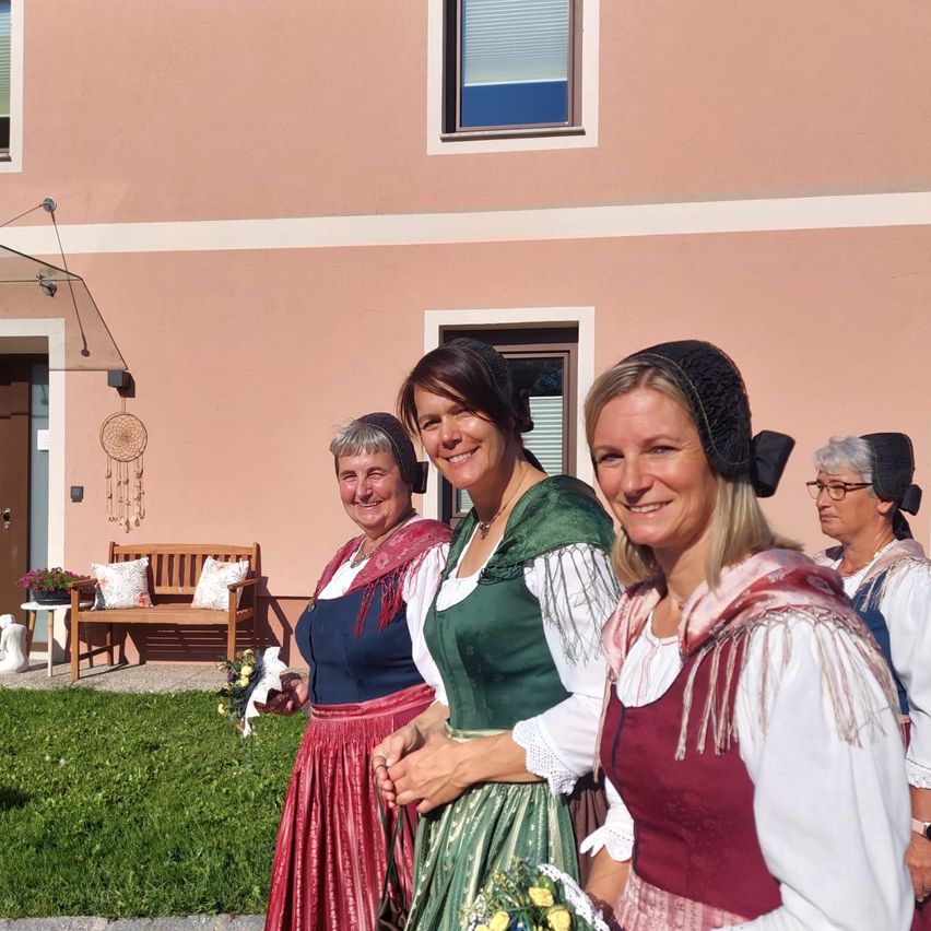 Vier Frauen in traditionellen Kostümen lächeln für ein Foto vor einem rosa Gebäude, mit einer Bank und Blumen im Hintergrund.