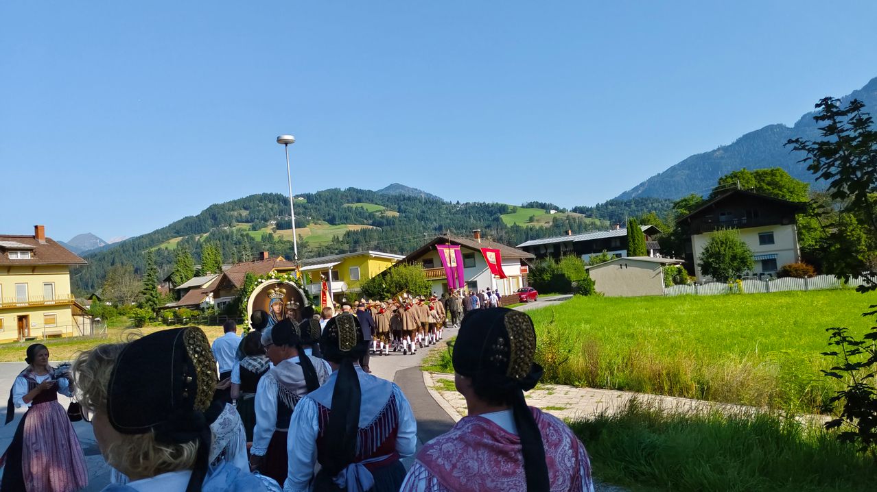 Eine Gruppe von Menschen in traditioneller Kleidung geht einen Pfad entlang, mit Bergen und Häusern im Hintergrund.