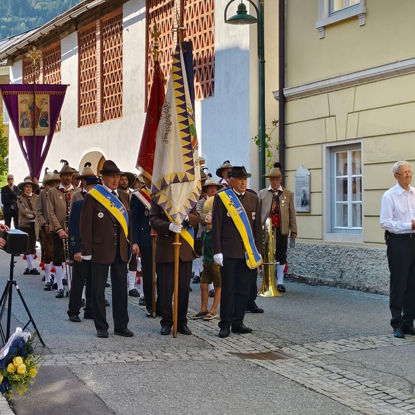 Eine Gruppe von Männern in Uniformen und Hüten steht vor einem Gebäude. Einige halten Fahnen und ein Blechblasinstrument.