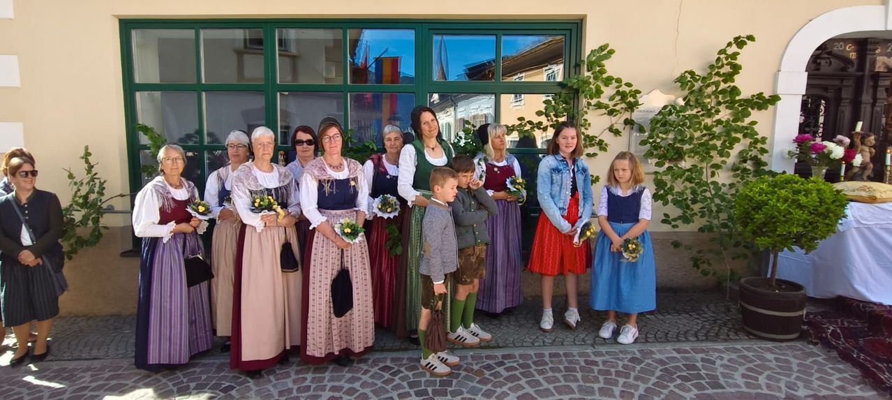 A group of adults and children in traditional attire stands in front of a building with a green-framed window, holding flowers. The children are wearing sneakers and the women are in traditional dresses.