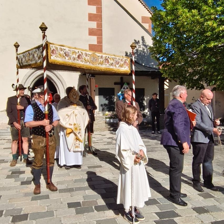 Several people in traditional attire stand in front of a church. A priest holds a child's hand, and others hold poles with ornate designs. A tree and a building are in the background.