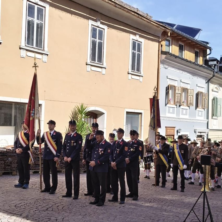 A uniformed group stands in a city square. They are holding flags and some wear hats. Behind them are buildings and people, some with musical instruments.