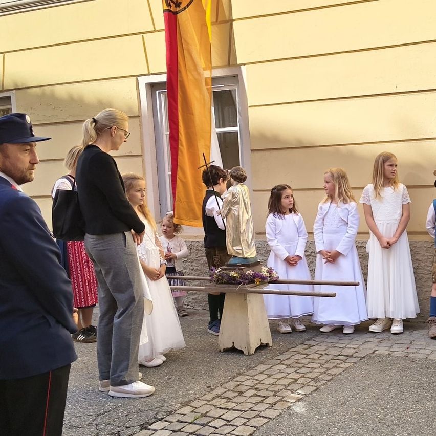 A group of young girls in white dresses are standing outside with a statue. A woman in black stands near them, and a man in a blue uniform is nearby. A flag hangs behind them on a building.