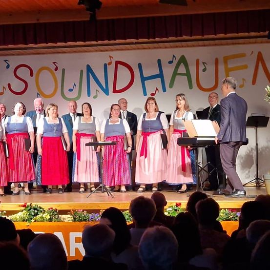 Ein Chor in traditionellen Kostümen singt auf der Bühne vor einem Banner mit der Aufschrift 'Soundhauen'. Dahinter steht ein Mann am Klavier, und das Publikum sitzt davor.