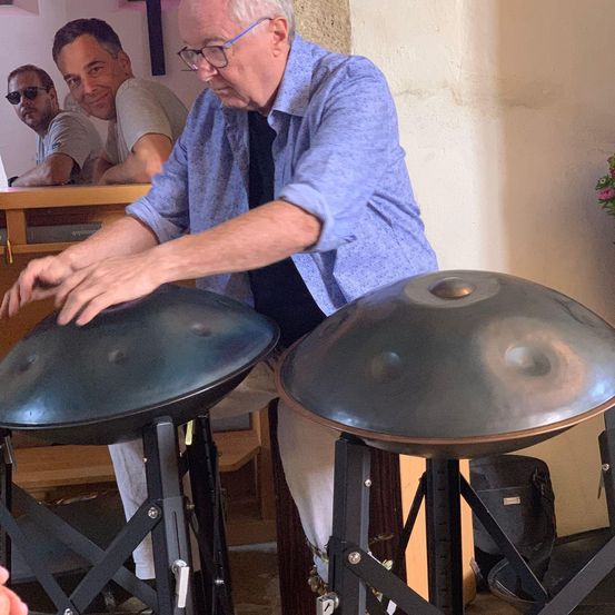 Ein älterer Mann spielt ein Handpan-Instrument, während er sitzt, und drei andere Männer beobachten ihn von hinten. Die Instrumente stehen auf Ständern, und eine Bank ist im Hintergrund zu sehen.