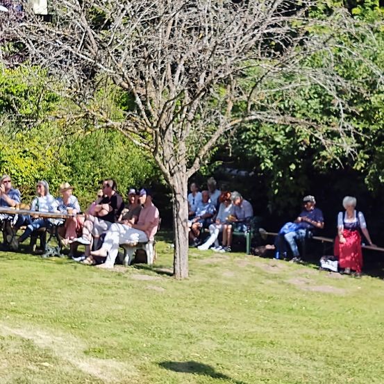 Menschen sitzen auf Bänken unter Bäumen in einem Garten. Einige tragen Hüte und Sonnenbrillen. Eine Frau in einem roten Kleid steht neben einer Bank.