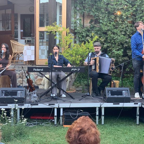 Eine Band spielt auf einer Bühne mit Geige, Akkordeon und Keyboard. Die Geigerin und Keyboarderin sind Frauen, der Akkordeonist ist ein Mann. Vor jedem steht ein Mikrofon.