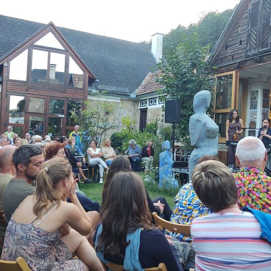 Eine Gruppe von Menschen sitzt auf Stühlen in einem Außenbereich und sieht sich eine Aufführung an. Vor ihnen steht eine blaue Statue. Einige tragen Hüte und Schuhe. Vor der Statue stehen zwei Menschen. Einer spielt Geige und der andere Flöte. Dahinter befinden sich Häuser mit Glasfenstern.