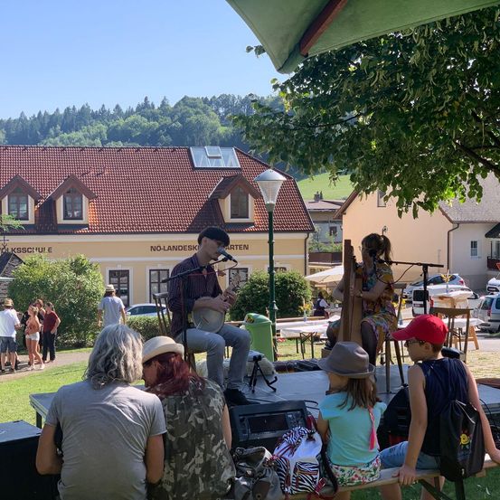 Eine kleine Band tritt im Freien auf, ein Mann spielt Banjo und eine Frau Harfe, umgeben von einem Publikum und einem Gebäude mit Solarpanelen.
