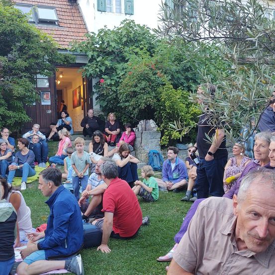 Eine Gruppe von Menschen sitzt auf einem Rasen, einige mit Turnschuhen, während ein Mann in der Nähe steht. Die Umgebung umfasst ein Haus mit einer offenen Tür und einer Person, die auf den Stufen sitzt. Im Hintergrund befinden sich Bäume und Pflanzen.