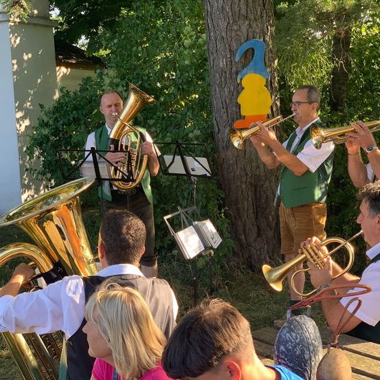 Eine Gruppe von Musikern, darunter Tuba- und Trompetenspieler, tritt im Freien in einer üppigen, grünen Umgebung auf. Das Publikum, bestehend aus mehreren Personen, sitzt vor ihnen.