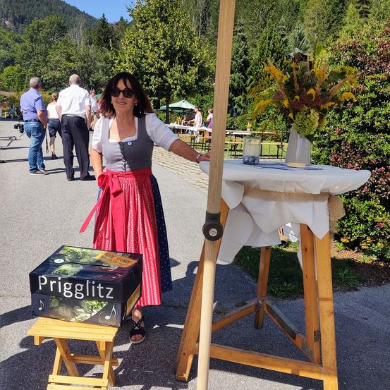 Eine Frau in traditioneller bayerischer Tracht steht neben einem Tisch mit einer Box namens Prigglitz und einer Blumenvase. Im Hintergrund gehen mehrere Personen in einem Park spazieren.