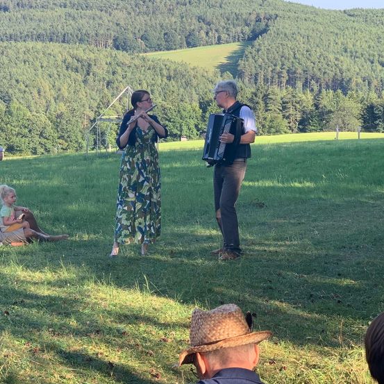 Eine Frau spielt Flöte und ein Mann spielt Akkordeon. Sie stehen in einem Feld, ein junges Kind sitzt in der Nähe. Dahinter ist ein Wald.