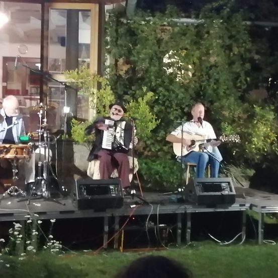 Drei Musiker spielen auf einer kleinen Bühne, einer spielt Akkordeon, ein anderer Gitarre und singt, und der dritte spielt Schlagzeug. Hinter ihnen stehen Pflanzen und ein Gebäude.