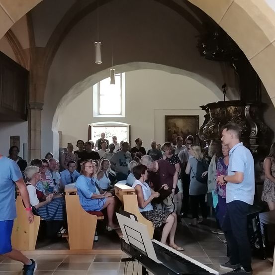 Eine Gruppe von Menschen sitzt auf Bänken in einer Kirche. Einige tragen Brillen und einer benutzt ein Handy. Ein Klavier mit Notenblättern steht auf dem Boden.